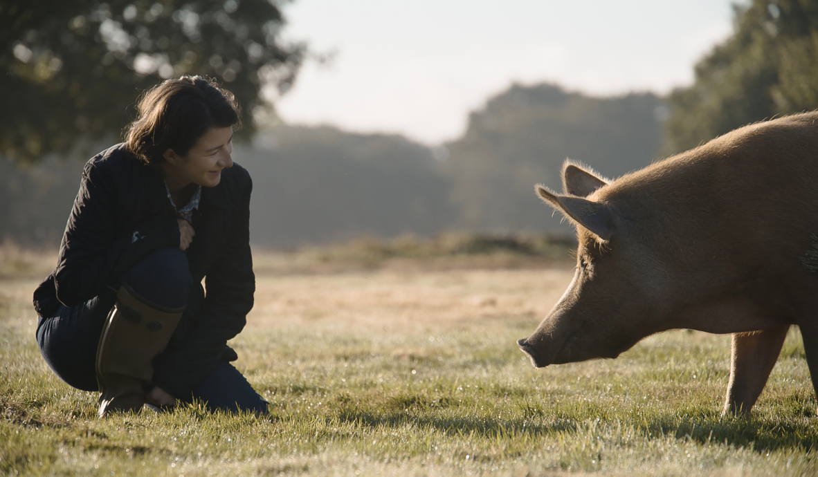 A white woman with short brown hair wearing a brown coat and jeans bending down in a grass field happily looking at a pig